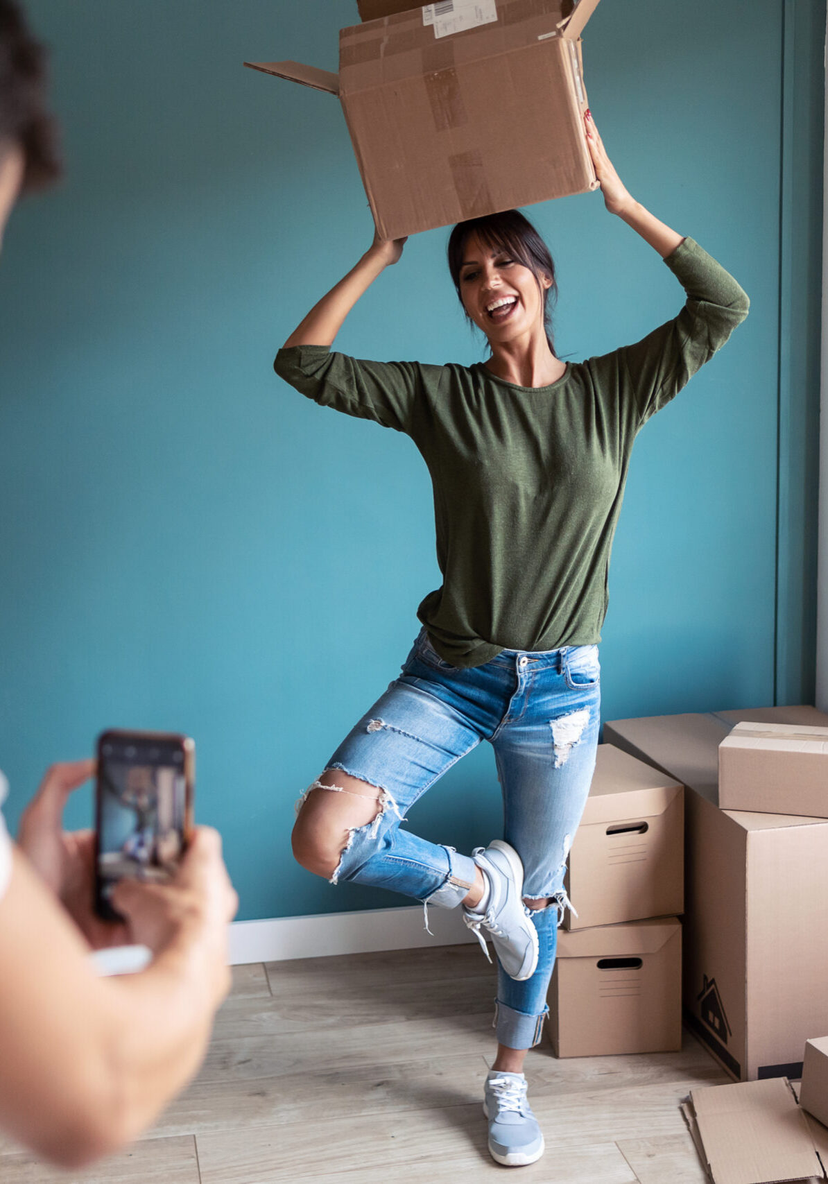 Shot of handsome man taking a photo with smart phone to his funny wife while she jokes with the moving cardboard boxes in the new house.